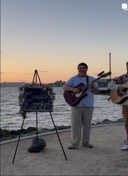 A band member playing guitar beside the water at sunset